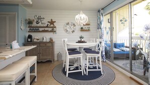 A dining area with a round table, white chairs, and a chandelier. There are shelves with decorative items and a clock on the wall. - 1BR Coastal Hideaway | Pool, Golf & Beach Nearby (Destin)