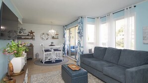A living room with a grey sofa, a dining area with a white table and chairs, and a kitchen area with a stove and decorative items. - 1BR Coastal Hideaway | Pool, Golf & Beach Nearby (Destin)