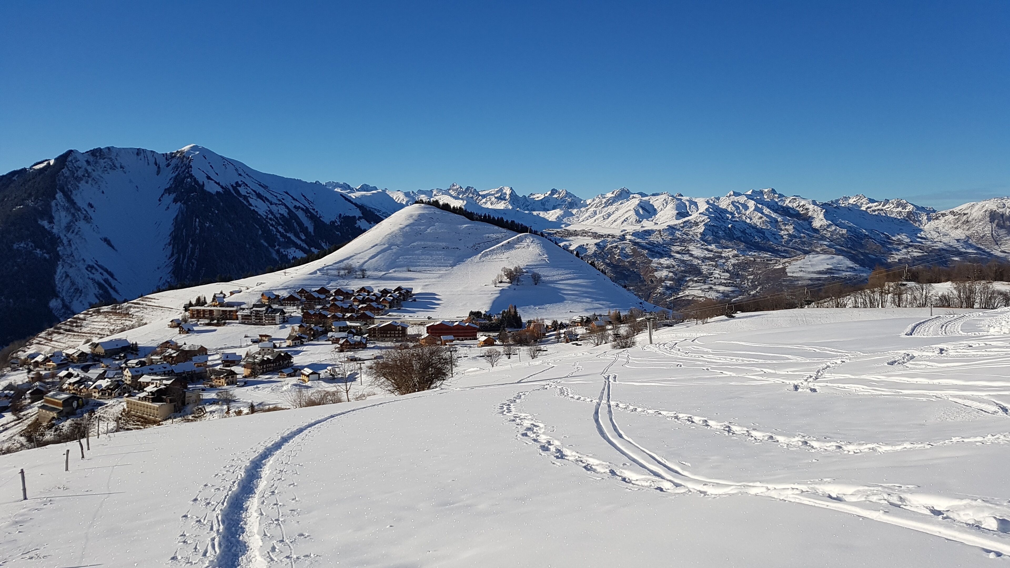 Un village de montagne enneigé, avec ses maisons traditionnelles en bois, une piste de ski et un ciel d’un bleu limpide.