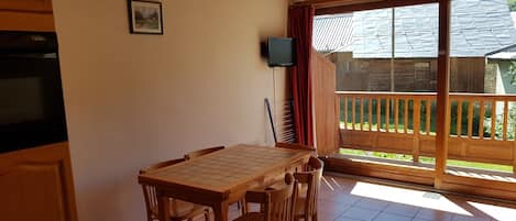 Un coin repas avec une table et des chaises en bois, un téléviseur fixé au mur, et une vue sur une terrasse en bois et la verdure environnante.