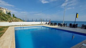 Ein rechteckiger Pool mit glasklar blauem Wasser, umgeben von einer gefliesten Liegefläche. Es gibt Sitzbereiche im Freien mit Tischen und Stühlen und Blick auf das Meer und die entfernten Hügel.