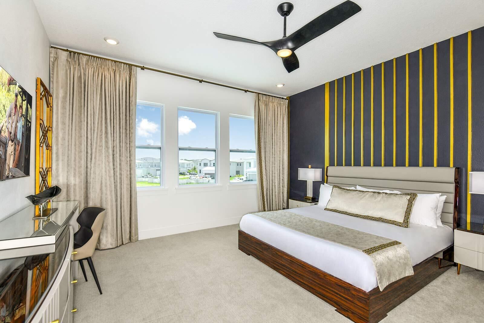 Une chambre d’hôtel moderne dotée d’un grand lit, d’un bureau avec une chaise, d’un ventilateur de plafond et de grandes fenêtres offrant une vue sur l’extérieur.