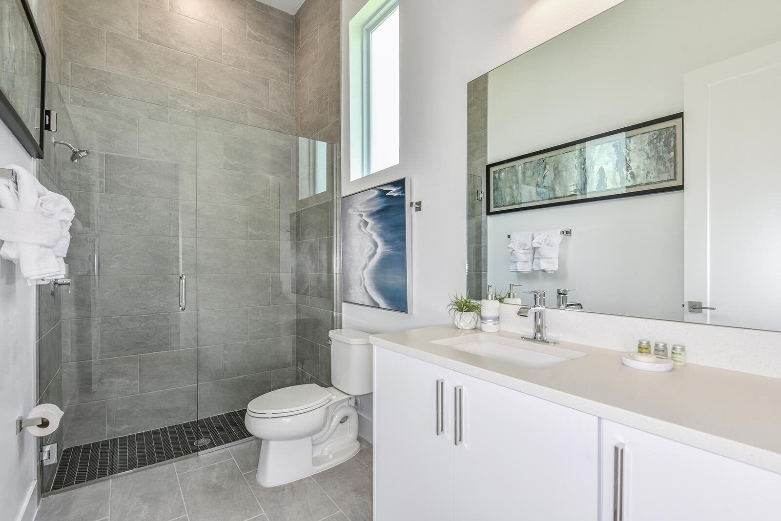 Une salle de bain moderne avec une douche à l’italienne, des toilettes, un lavabo doté d’un grand miroir et une œuvre d’art encadrée au-dessus du miroir.