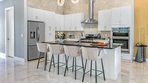 Une cuisine moderne avec des meubles blancs, un îlot central doté de tabourets de bar, des appareils en acier inoxydable et un éclairage suspendu.