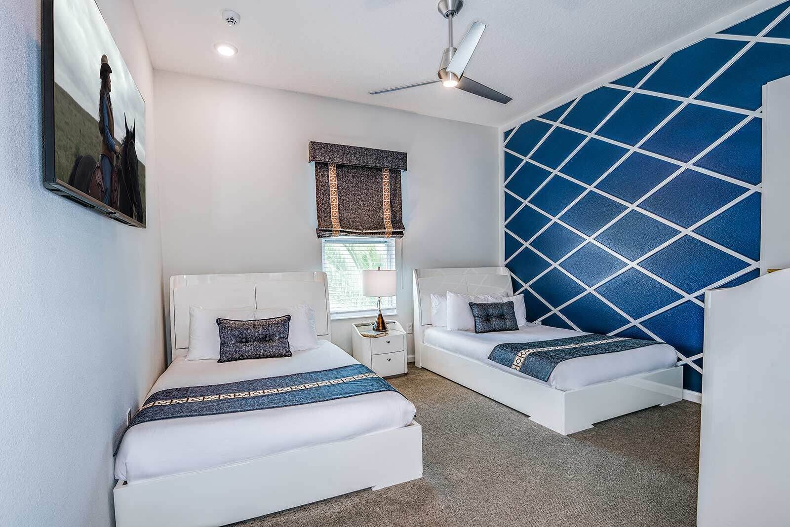 Un dormitorio con dos camas, ventilador de techo, ventana con persianas y un diseño geométrico azul en la pared.