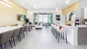 Une cuisine moderne dotée d’un îlot central, un coin repas avec une longue table et un espace de vie comprenant un canapé et un téléviseur.