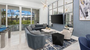 A modern living room with a pool table, a sectional sofa, a flat-screen TV, and a large window offering a view of a pool and greenery. - Stylish Home Near Disney w/ Secret Gaming RM, Pirate BR, Theater & Pool Table (Davenport)
