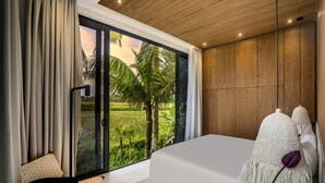 A modern bedroom with a large bed, a view of a lush garden, and a cozy seating area. - Green Flow Villa 16 (Ubud)