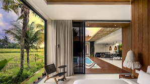 A modern bedroom with a large bed, a view of a pool and outdoor seating, and a glass door leading to a balcony. - Green Flow Villa 16 (Ubud)