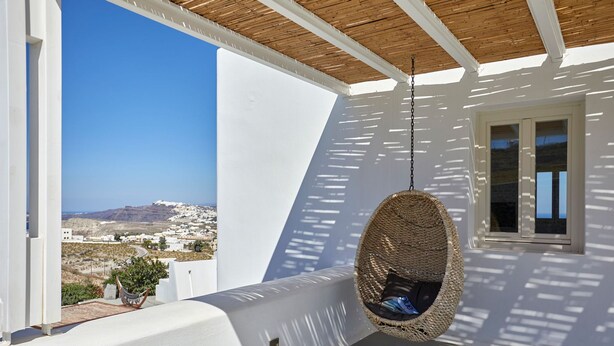 A white balcony with a hanging chair, a window, and a view of a coastal town. - Exclusive One Bedroom Villa - White Co (Santorini)