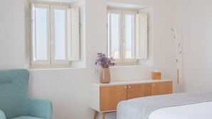 A bedroom with a bed, a blue chair, a wooden dresser, and two windows. - Exclusive One Bedroom Villa - White Co (Santorini)