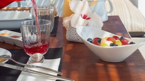 Una mesa con una copa de líquido rojo, una jarrón, una copa de líquido amarillo, un cuenco de fruta y un jarrón blanco con flores.