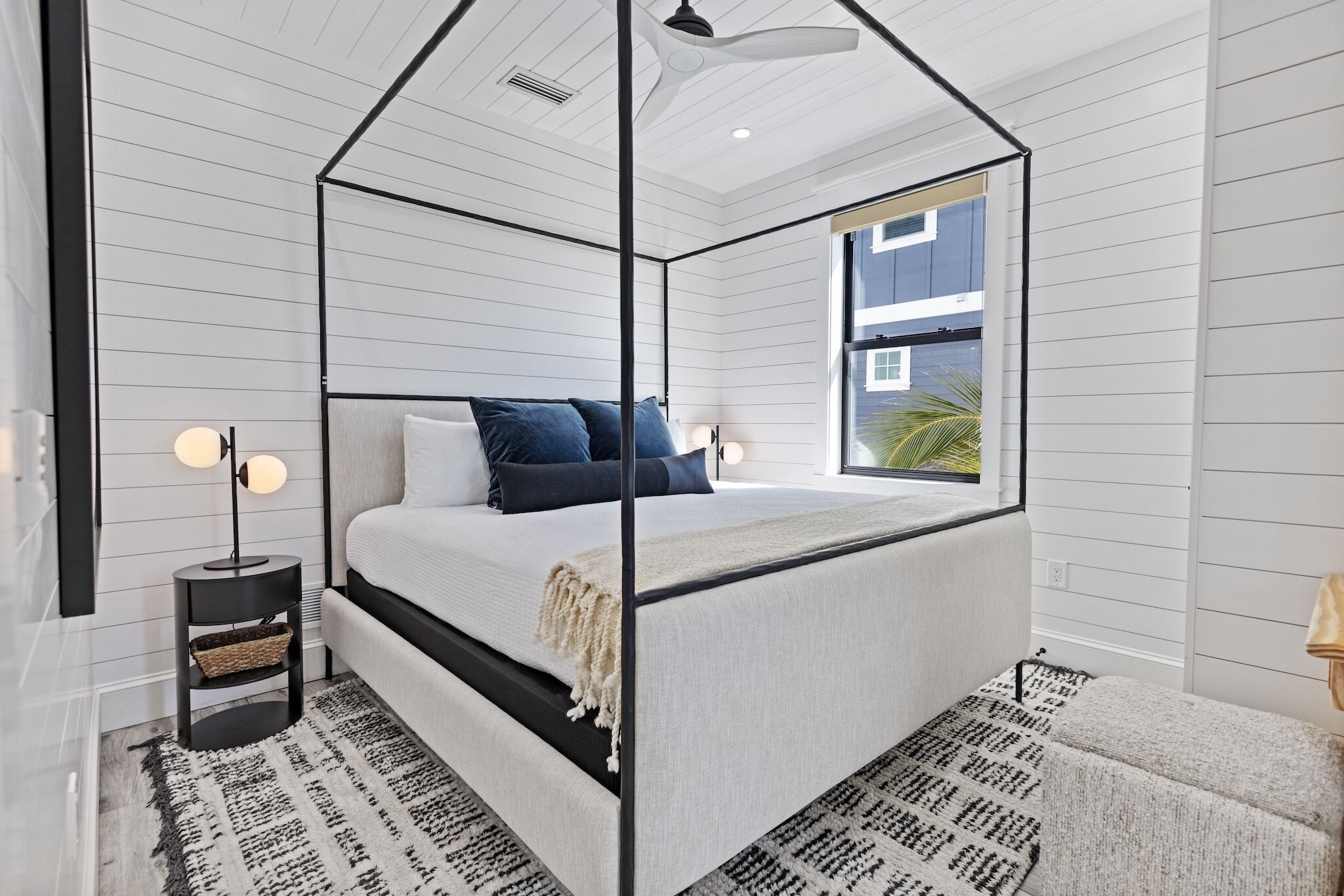 A modern bedroom with a four-poster bed, a small side table, and a large window.