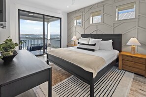 Ett modernt sovrum med en stor säng, två sängbord, ett litet bord, en krukväxt, en balkong med utsikt över vattnet och en vägg med ett geometriskt mönster.