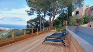 Une terrasse en bois avec des chaises longues, donnant sur une piscine et offrant une vue panoramique sur la mer.