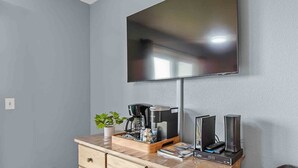 Un salon moderne avec un téléviseur à écran plat fixé au mur, une table basse en bois sur laquelle se trouve une machine à café, une plante et une horloge numérique affichant l’heure.