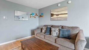 Un salon avec un canapé gris, deux coussins à motifs, une table basse en bois, un miroir et deux œuvres d’art accrochées au mur.