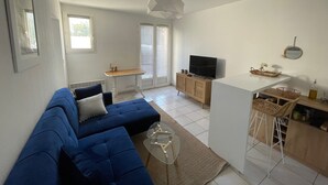 Una sala de estar moderna con un sofá azul, una mesa de centro de vidrio, un televisor sobre un mueble de madera, y una zona de cocina con un taburete de bar.