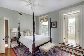 A four-poster bed with a white bedspread, a wooden bench, a patterned rug, and a ceiling fan. - Liberty House, Historic Home | Centrally Located (Savannah)