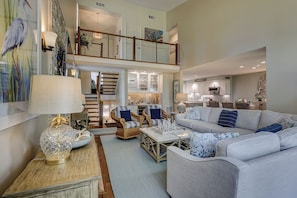 A spacious living area with a high ceiling, a staircase leading to an upper level, and a kitchen area visible in the background.