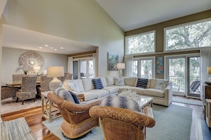 A spacious living room with a large sectional sofa, wicker chairs, a coffee table, and large windows offering a view of trees.