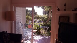 A living room with a sliding glass door leading to a patio. There is a TV on the right, a lamp on the left, and a chair near the door. - Apartamento con Encanto al Lado de la Playa El Golfet (Calella de Palafrugell)