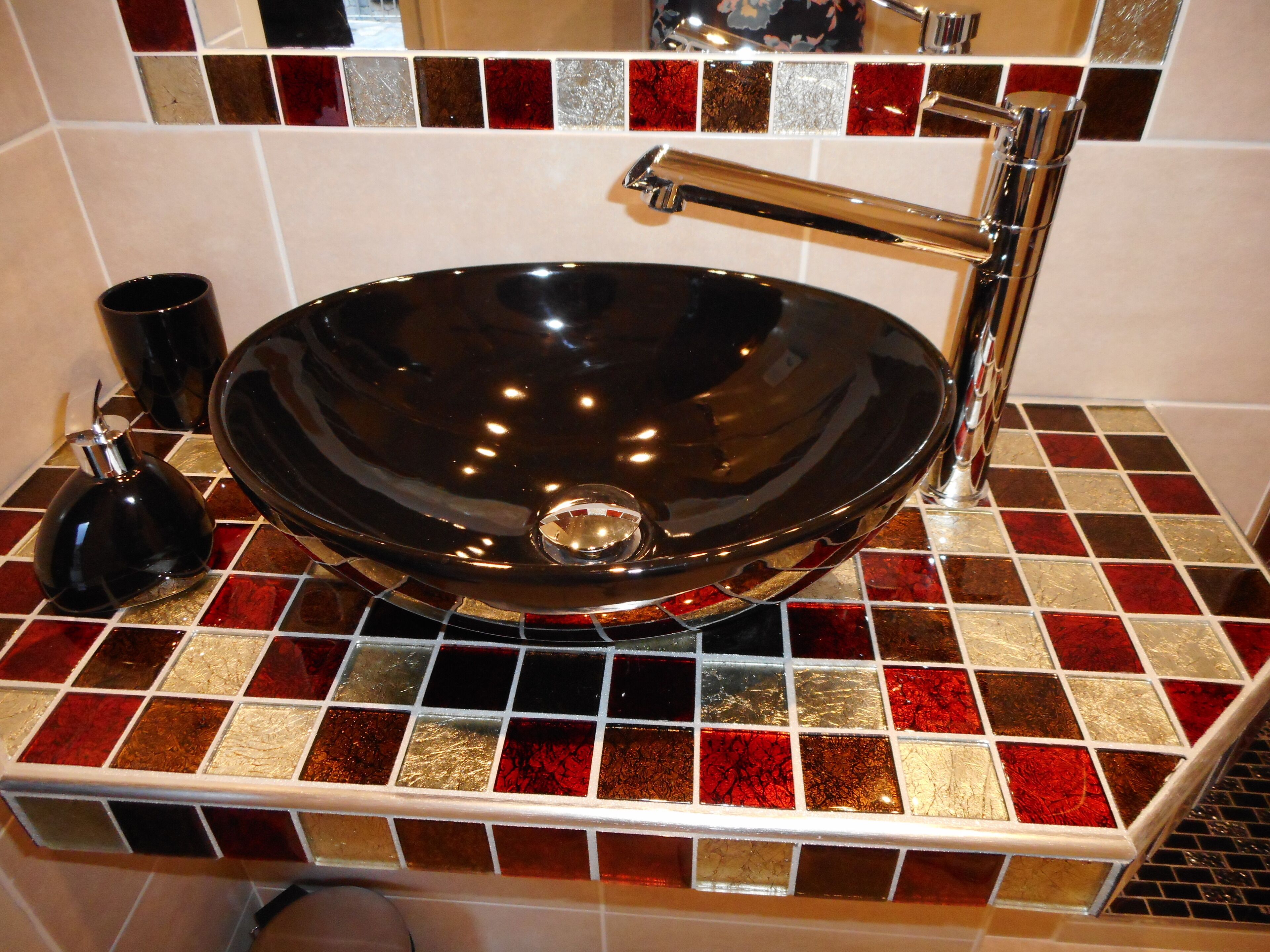 A bathroom with a black vessel sink, chrome faucet, and a tiled backsplash with red, gold, and white tiles.