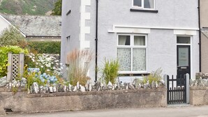 Ein zweistöckiges Haus mit weißer Fassade, einem schwarzen Dach und einer kleinen Veranda mit Steinwand und Pflanzen.