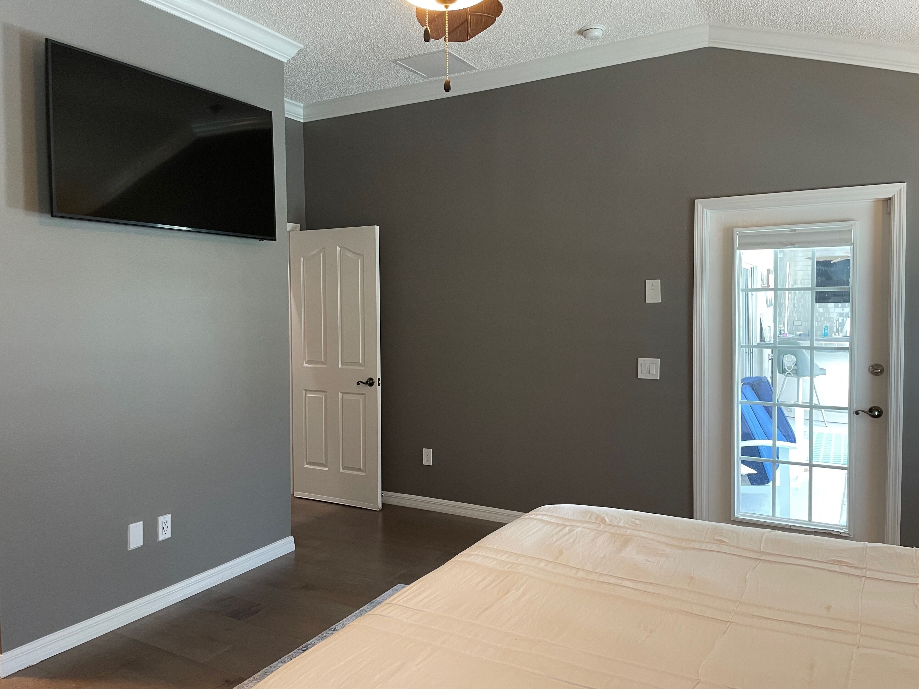 A bedroom with a flat-screen TV mounted on the wall, a door, and a window with a view of the outside.