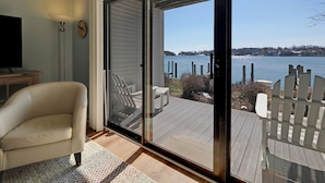 A room with a large sliding glass door leading to a deck with a view of a lake and distant buildings. - First-Floor Lakefront Gem | Downtown Charlevoix | Pool, Hot Tub, Sauna (Charlevoix)