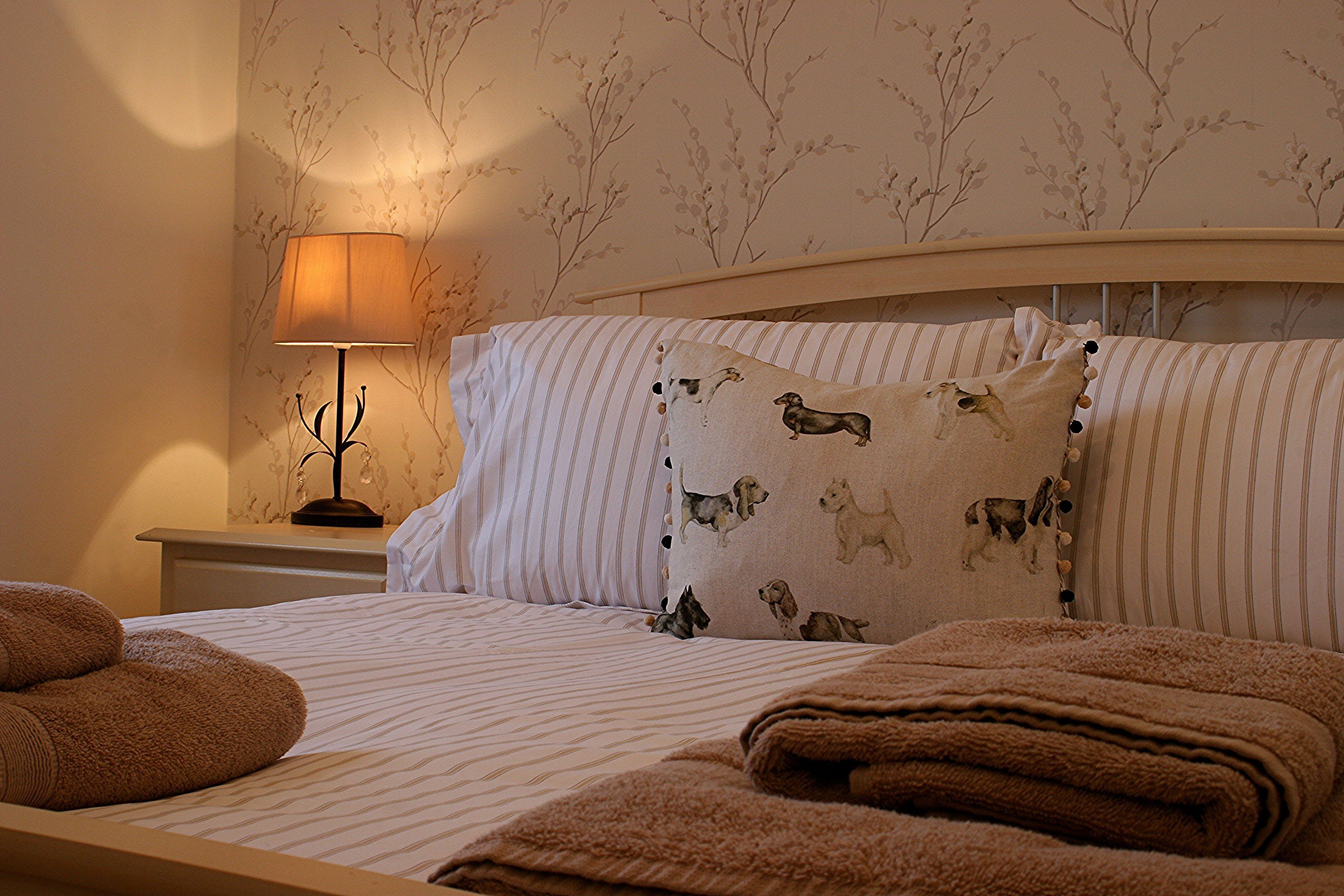 A neatly made bed with striped bedding and a pillow featuring dog illustrations, a bedside lamp, and a wall with a floral pattern.