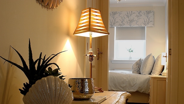 A bedroom with a bed, a bedside table with a lamp, a potted plant, and a window with a view of trees. - Linden Apartment -  an apartment that sleeps 4 guests  in 2 bedrooms (Stirling)