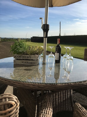 Una mesa redonda de vidrio con una sombrilla metálica, una botella de vino y tres copas de vino.