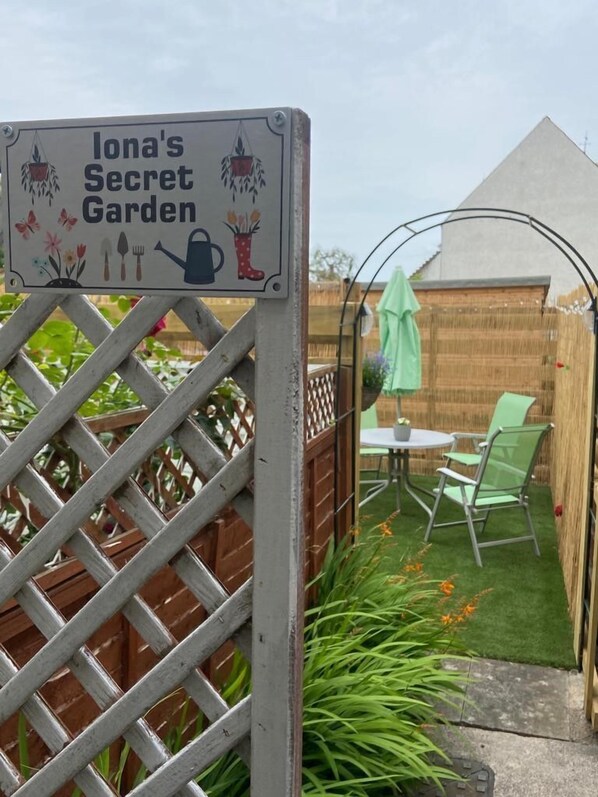 Un letrero de jardín para "Iona's Secret Garden" con un cerco de celosía de madera, una pequeña mesa con sillas y un paraguas verde.