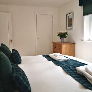 Una cama bien hecha con una manta escocesa, una mesita de noche de madera con un jarrón de flores y una puerta con manija de latón.