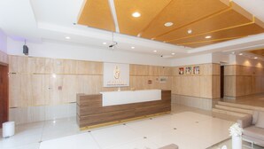 A hotel reception area with a wooden reception desk, a white wall with a logo, and a clean, tiled floor.