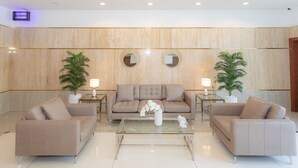 A modern lobby with a beige sofa set, a glass coffee table, and two side tables with lamps and potted plants.