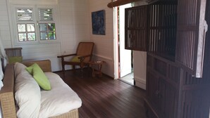 A cozy room with a wooden floor, a wicker sofa with cushions, a wooden chair, a small table with a book, and a large wooden door. - Charming bungalow for 2 with private Jacuzzi (Saint-François)