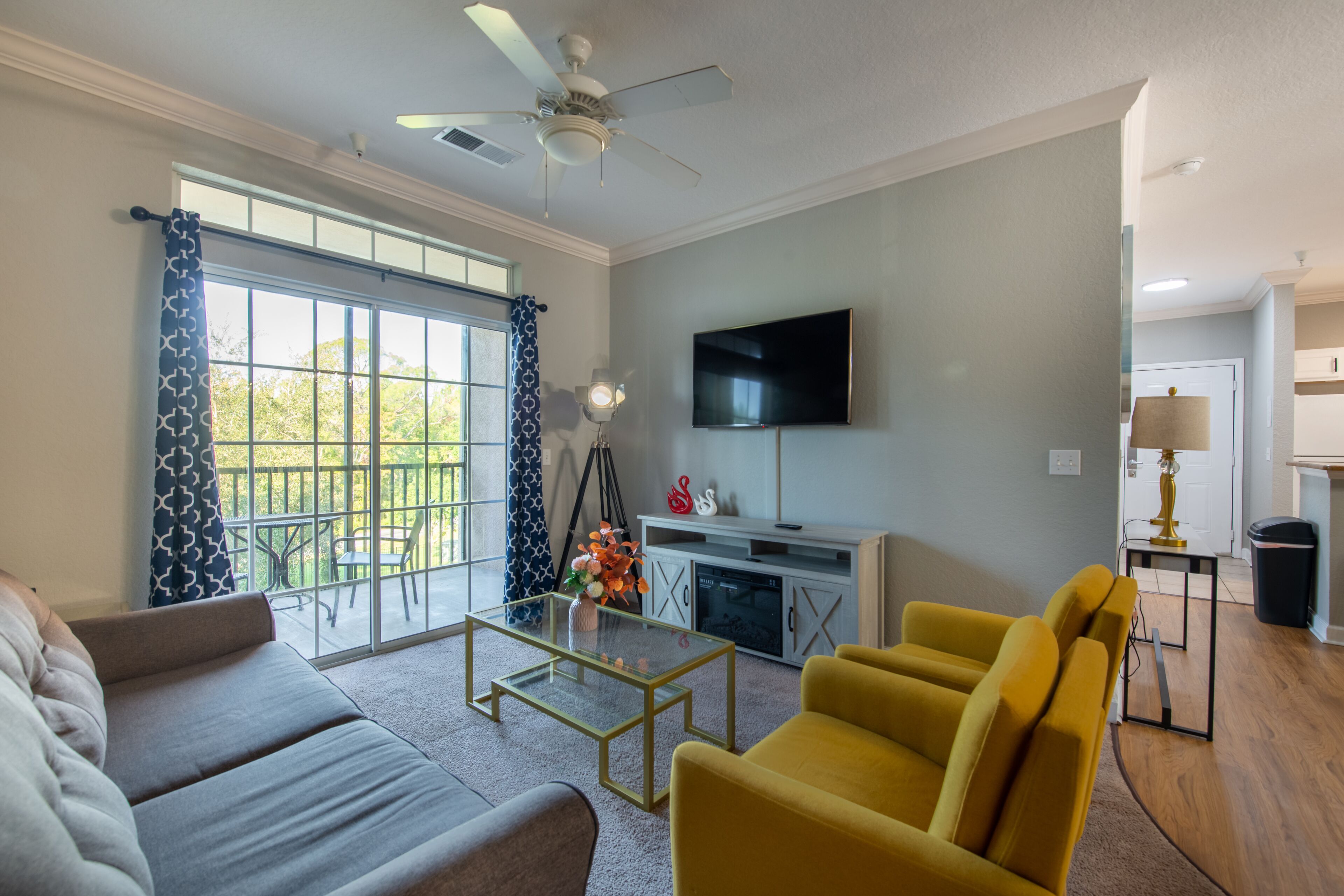 A living room with a sofa, yellow armchair, glass coffee table, and a flat-screen TV mounted on the wall.