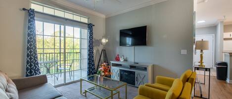 A living room with a sofa, yellow armchair, glass coffee table, and a flat-screen TV mounted on the wall.
