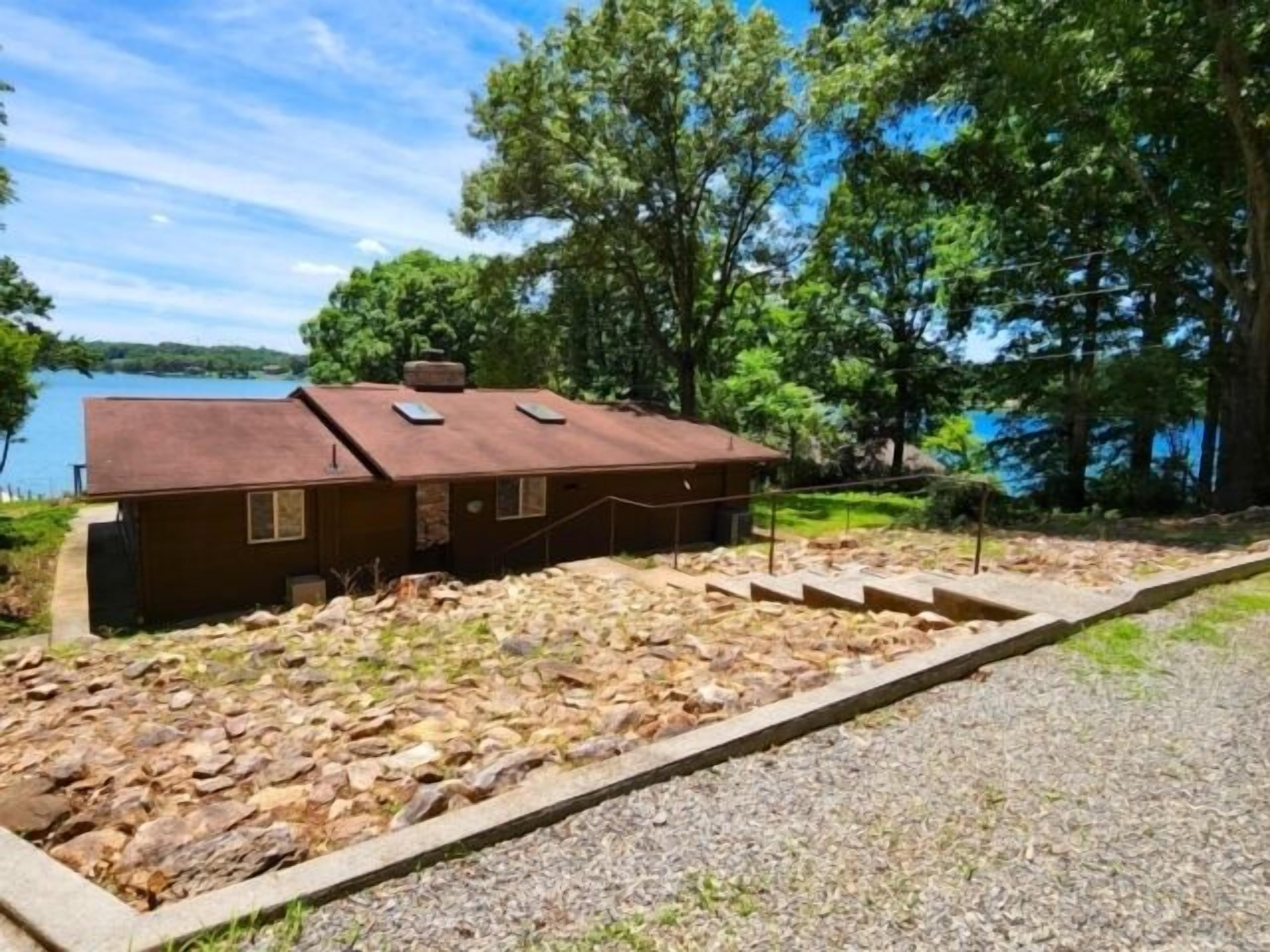 A cabin with a stone foundation, a gravel driveway, and a view of a lake.