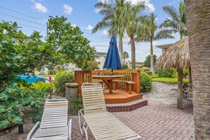 A patio area with two lounge chairs, a wooden deck, and a blue umbrella. - Friendly Native Unit 507- Fun and relaxation on vibrant St Pete Beach! (St Pete Beach)