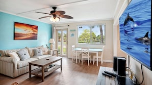 A living room with a striped sofa, a coffee table, and a dining area with white chairs. There is a ceiling fan, a painting of a sunset, and a large aquarium with dolphins. - Friendly Native Unit 507- Fun and relaxation on vibrant St Pete Beach! (St Pete Beach)