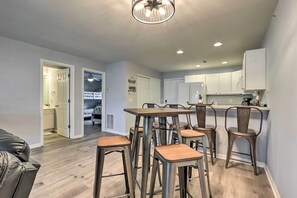 A modern kitchen with white cabinets, a dining area with metal stools, and a visible doorway leading to another room.