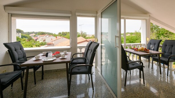 A balcony with a table and chairs, a glass door, and a view of a residential area. - Apartment SoMi 2 (Poreč)