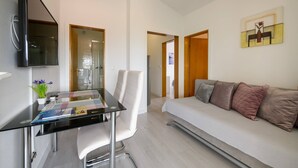 A modern room with a glass desk, white chairs, a sofa with multiple cushions, a flat-screen TV, and a painting on the wall. - Apartment SoMi 2 (Poreč)