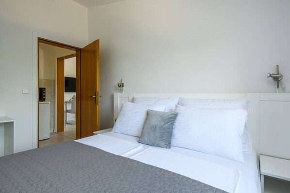Une chambre à coucher avec un lit, des murs blancs, une porte en bois et une salle de bain visible à travers une porte ouverte.