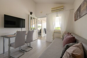 Une chambre moderne dotée d’une télévision à écran plat, d’une table à manger avec des chaises, d’un canapé avec des coussins et d’une grande fenêtre avec des rideaux.