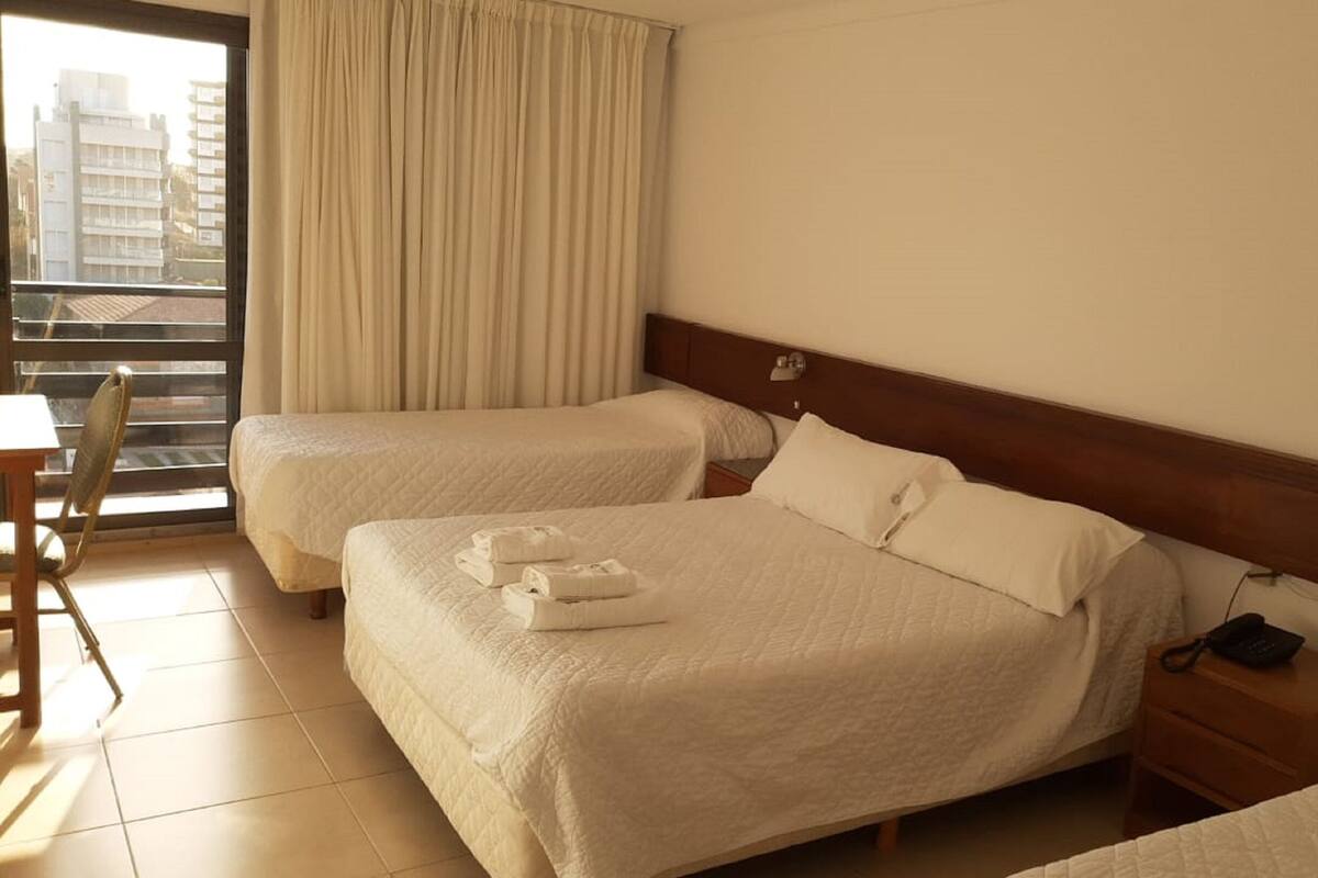A hotel room with two beds, a chair, a nightstand, and a view of buildings through the window.
