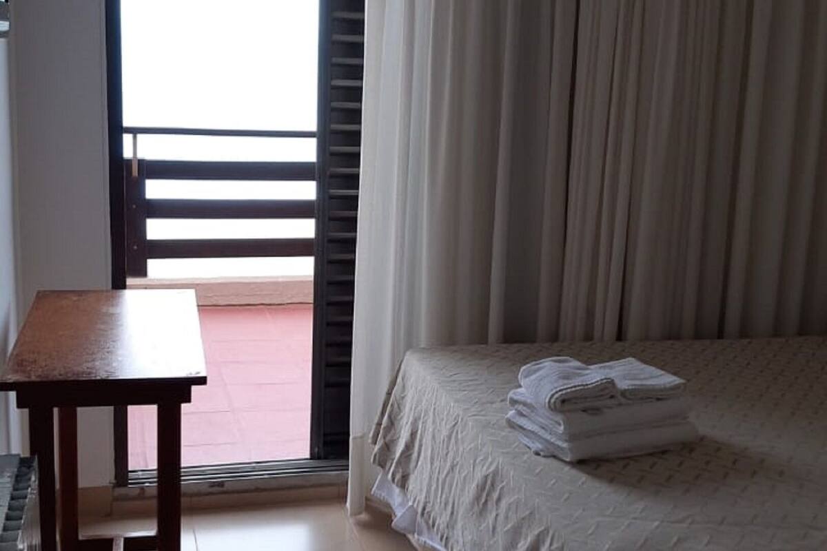 A bedroom with a bed, a table, a fan, and a view of the outside.